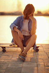 A young beautiful girl thoughtfully sitting on a skateboard at the dock and enjoying the sunset on the sea. Summer, sea, vacation
