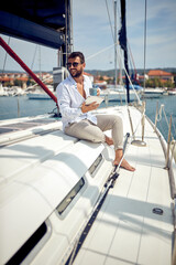 A young man with a tablet is sitting on the yacht and enjoying a coffee and beautiful scenery of the dock on the seaside. Summer, sea, vacation