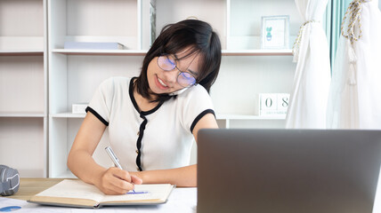 Asian female worker talking on phone and working on laptop with smiling face, Office staff work and telephone conversations, Take notes for reminders, Business conversation over the phone.