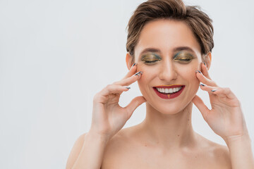 satisfied young woman with shimmery eye makeup touching cheeks isolated on grey.