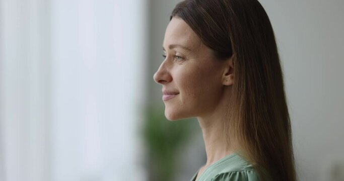 Close up shot, profile of dreamy smiling Caucasian woman standing in living room, look out window, feels happy while visualizing good future, dreaming about opportunities, starting new day. Daydreams