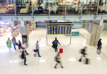 Blurred people walking in an airport hall next to a standing woman reading information from the...