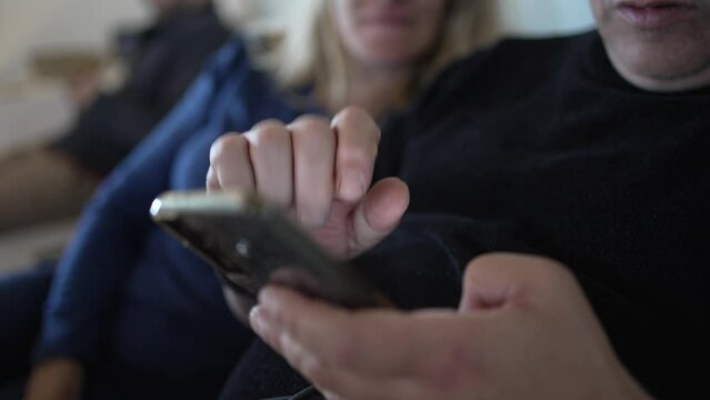 couple on a sofa looking at a mobile phone in the background another guy out of focus watching TV, detail of hands touching the phone and configuring applications together.