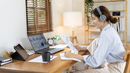 Asian female college student wearing headphones watching live performance or video call teacher teaching on laptop, Conversations with teachers and classmates, Online learning, Study at your own home.