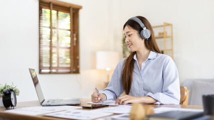 Asian female college student wearing headphones watching live performance or video call teacher teaching on laptop, Conversations with teachers and classmates, Online learning, Study at your own home.