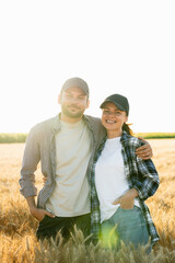 A couple of farmers in plaid shirts and caps stand embracing on agricultural field of wheat at sunset	