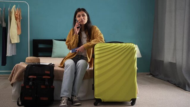 Happy woman sitting near suitcase on bed, speaking on smartphone, booking travel tour, making hotel reservation or buying tickets from home. Travel