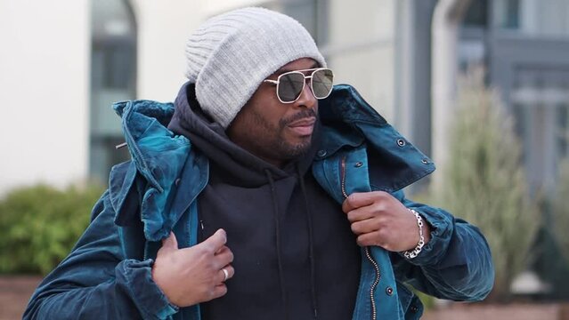 Portrait of handsome African American man walking street, wearing stylish outfit parka coat, knitted hat and sunglasses. Street style fashion male model