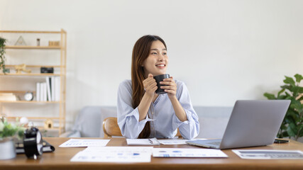 Beautiful Asian woman working on laptop and sipping coffee with smiling face in her home, Creating happiness at work with a smile, Freelancer working at home happily, Work from home.