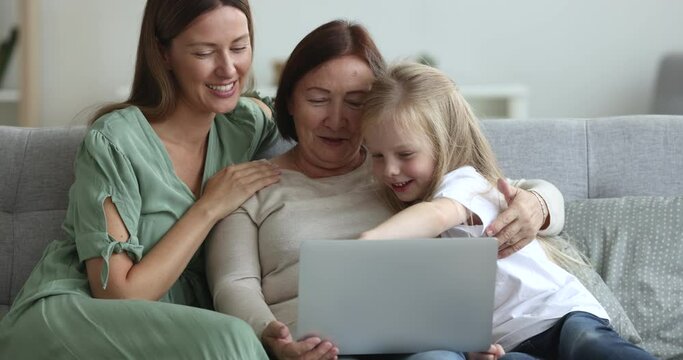 Little girl with mom and grandma sit on sofa watch on-line content on laptop, spend weekend together at home, shopping, order services, use educational app for kids, choose goods, plan joint vacation