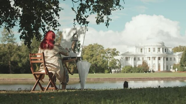 Back view of red haired young female artist sitting by river in beautiful summer park painting landscape