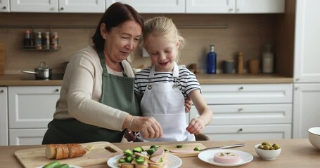 Older grandma and little grandkid wear aprons cooking together in the kitchen, loving grandmother teach cute preschooler girl to cook, prepare sandwiches for breakfast, kid help to granny with chores - Powered by Adobe