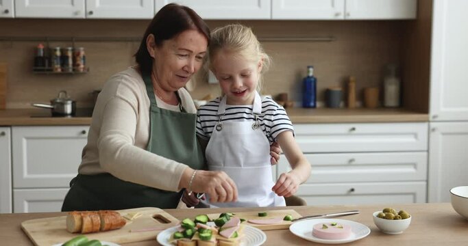 Older grandma and little grandkid wear aprons cooking together in the kitchen, loving grandmother teach cute preschooler girl to cook, prepare sandwiches for breakfast, kid help to granny with chores