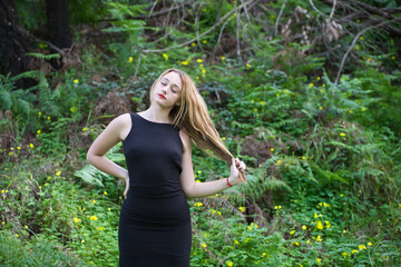 Young, beautiful blonde woman dressed in black walks through the forest in different postures and expressions. In the background ferns and yellow flowers. Concept expressions in nature.