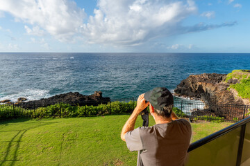 Mature Caucasian man watching a boat far on the ocean with binoculars from a balcony, lanai, with...
