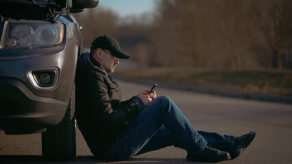 Upset man using smartphone application for asking help repair service because his car is broken. Driver sitting near pick up with damaged engine. Male on road with problem emergency insurance auto