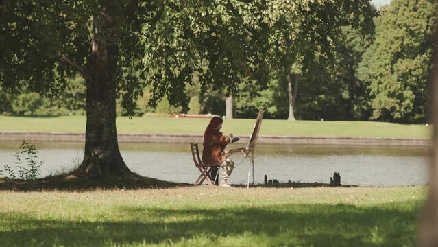 Wide shot of young female artist sitting on sunlit green grass in park by water painting on canvas