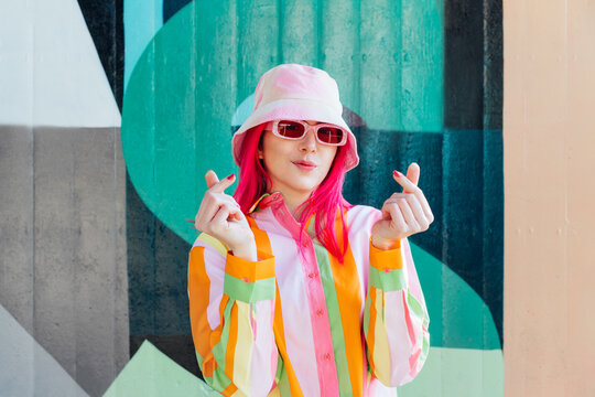 Vanilla Girl. Kawaii vibes. Candy colors design. Young woman with pink hair and sun glasses in bucket hat and multicolor shirt making Finger Heart sign on wall background. Love fashion concept