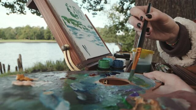 Cropped shot of unrecognizable female artist painting landscape on canvas while sitting by water outdoors in park
