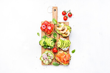 Avocado sandwiches or toasts with salmon, shrimps, tomatoes, cucumbers, soft cheese and spinach, cashew nuts and sesame seeds on wooden cutting board, white table background, top view