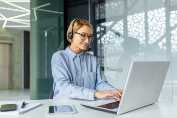 Successful businesswoman with video call headset working inside modern office, online customer support worker using laptop typing on keyboard, successful woman smiling satisfied with work.