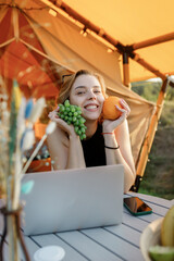 Smiling Woman freelancer holding grape and orange while working on laptop in cozy glamping tent in a sunny day. Luxury camping tent for outdoor summer holiday and vacation. Lifestyle concept