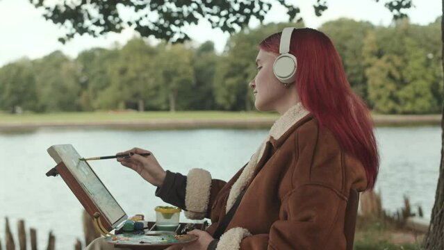 Medium side view shot of Caucasian teenage girl with colored pink hair enjoying music in headphones while painting landscape on canvas spending leisure time alone in park by water