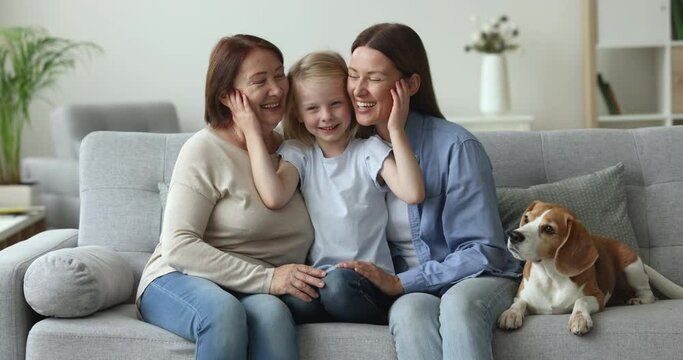 Happy three generations of women sit on sofa in living room, look at camera, laugh, pose for family picture together with cute dog, show tenderness and care for each other, enjoy warm relationships