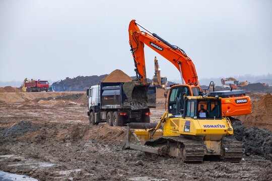 Ust-Luga, Leningrad oblast, Russia - November 16, 2021: Excavator Doosan, bulldozer Komatsu and dump trucks on construction site. Rainy day, muddy road