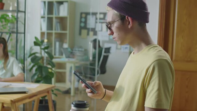 Medium shot of young gen Z guy in casual outfit, glasses and beanie hat holding to go coffee cup and scrolling on smartphone while standing in office