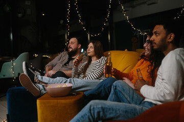 A company of multiracial friends watching a movie sitting on soft bean bags and drinking beer.