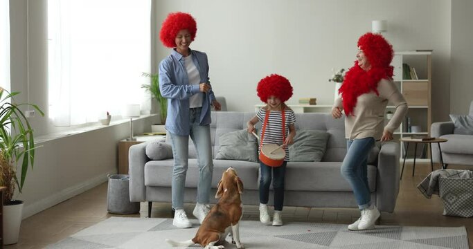 Multi-generational family wear red wigs marching to the sound of drum play by little cute girl having fun together in cozy living room with barking beagle dog. Family weekend fun and pastime. Hobby
