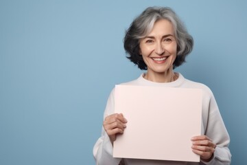 Naklejka premium Portrait of smiling senior woman holding blank sheet of paper on blue background