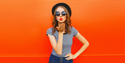 Portrait of beautiful young woman blowing her lips sending sweet air kiss wearing black round hat on orange background