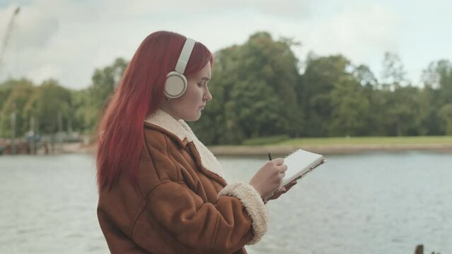 Medium shot of young Caucasian female artist with bright red hair enjoying music in headphones while drawing landscape in sketchbook outdoors in park