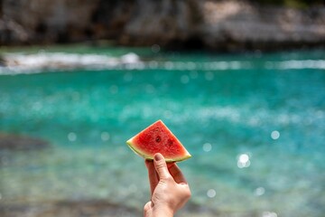 Fototapeta na wymiar A hand holding a piece of watermelon with turquoise sea in the background