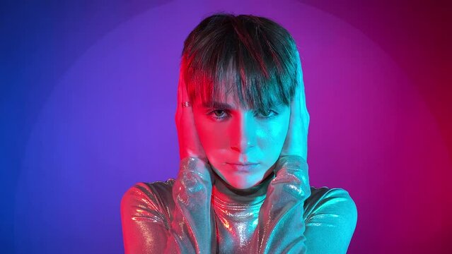 Portrait young adult woman covering ears hands, showing not listening gesture, looking camera neon illumination indoors. Studio shot art colorful lighting. Female short hair wearing sparkling clothing