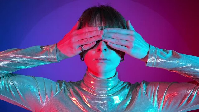 Portrait young woman covering eyes with hands, showing see no evil gesture, looking camera in neon illumination indoors. Studio shot art colorful lighting. Female short hair wearing sparkling clothing