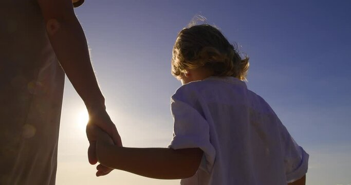 A child and a woman walk, holding hands, view from behind. Woman took child from orphanage and took him for a walk to get closer. Little boy tries to get used to his new adoptive mom while they walk.