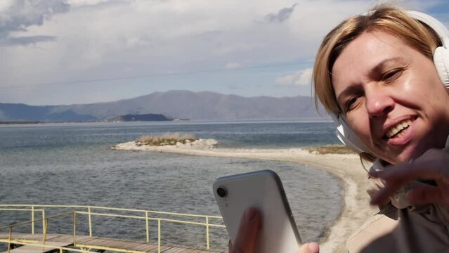 Cheerful young adult woman talking via video call using mobile phone and wireless white headphones while on the shore of a beautiful mountain lake or sea on a sunny but windy day on vacation