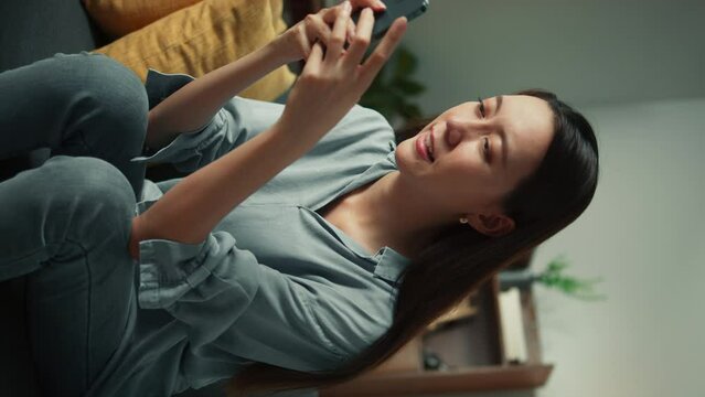Happy beautiful Asian woman sit on sofa hand holding mobile phone chatting and playing social media. Female looking at smartphone cellphone browsing internet on sofa home living room. Vertical Shot