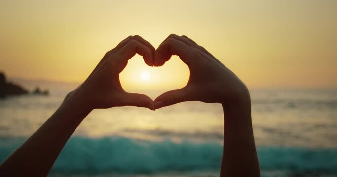 Woman on beach making heart with hands at sunset light on ocean. Sign of love close-up in slow-motion.