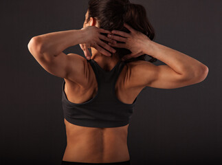 Sporty muscular woman with ponytail doing stretching workout of the shoulders, blades in sport bra, holding hands behind the head  on dark grey background with empty space. Closeup portrait.