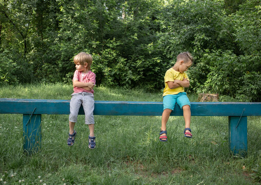 Two brothers or boys friends in casual clothes are sitting on bench offendedly turned away in different directions, sad and gloomy. Offended, dissatisfied, do not want to agree with each other.
