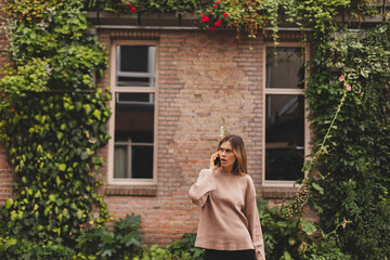 Portrait of unhappy frustrated blonde woman outdoor talking on phone. Woman wear beige t-shirt and look at side with open mouth. Brick and green trees background.