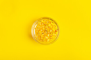 Vitamin d3 yellow soft gel capsules in small glass bowl on bright yellow background from above.