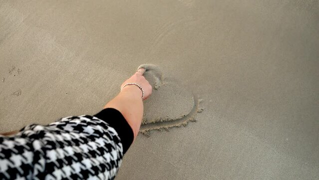 Closeup of female hand drawing a heart shape of wet sand at sea beach. Concept of loving travel, tips, journey and tourism