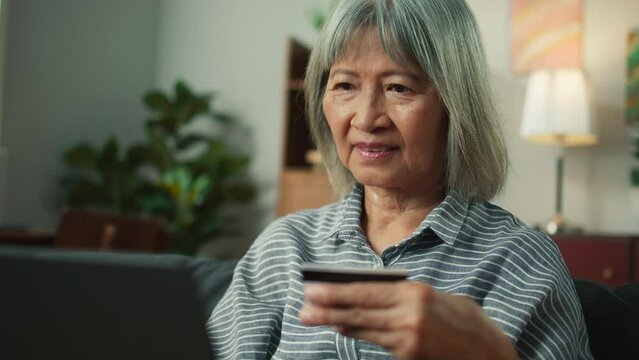 Attractive Asian elderly woman shopping online using laptop computer holding credit card purchase via e-banking. Beautiful old grandma making online payment through e-commerce platform in living room.