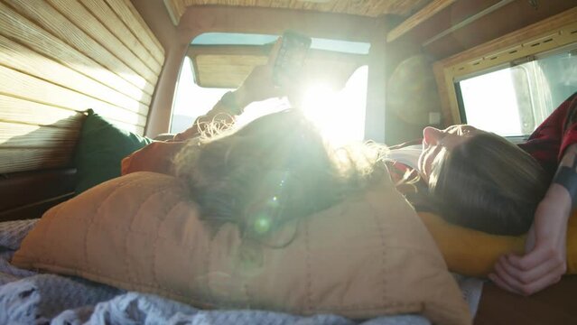 Young caucasian couple lying on back at a bed in small cozy camper van. Attractive hipster man shoving something on his phone to his girlfriend. Romantic road trip, outdoor weekend, bright daylight