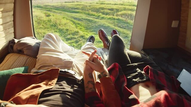 View of young caucasian couple lying on back at a bed in small cozy camper van and holding hands. Close up, warm colors. Romantic road trip, hipster pair in love on outdoor vacation